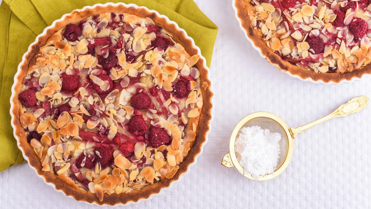 Frangipane-Tarte mit Rhabarber und Himbeeren. Daneben in goldenes Sieb mit Puderzucker.