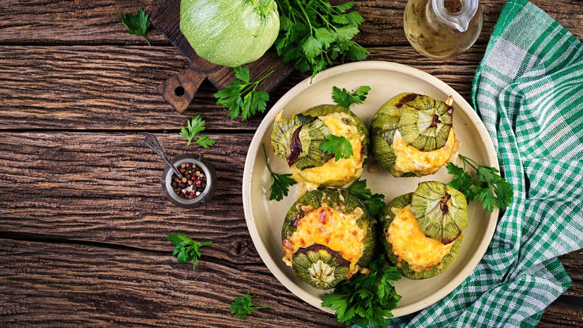 Vier gefüllte Zucchini mit Soja-Hack und Käse und Petersilie auf einem Teller, Holzuntergrund und Zutaten drum herum.