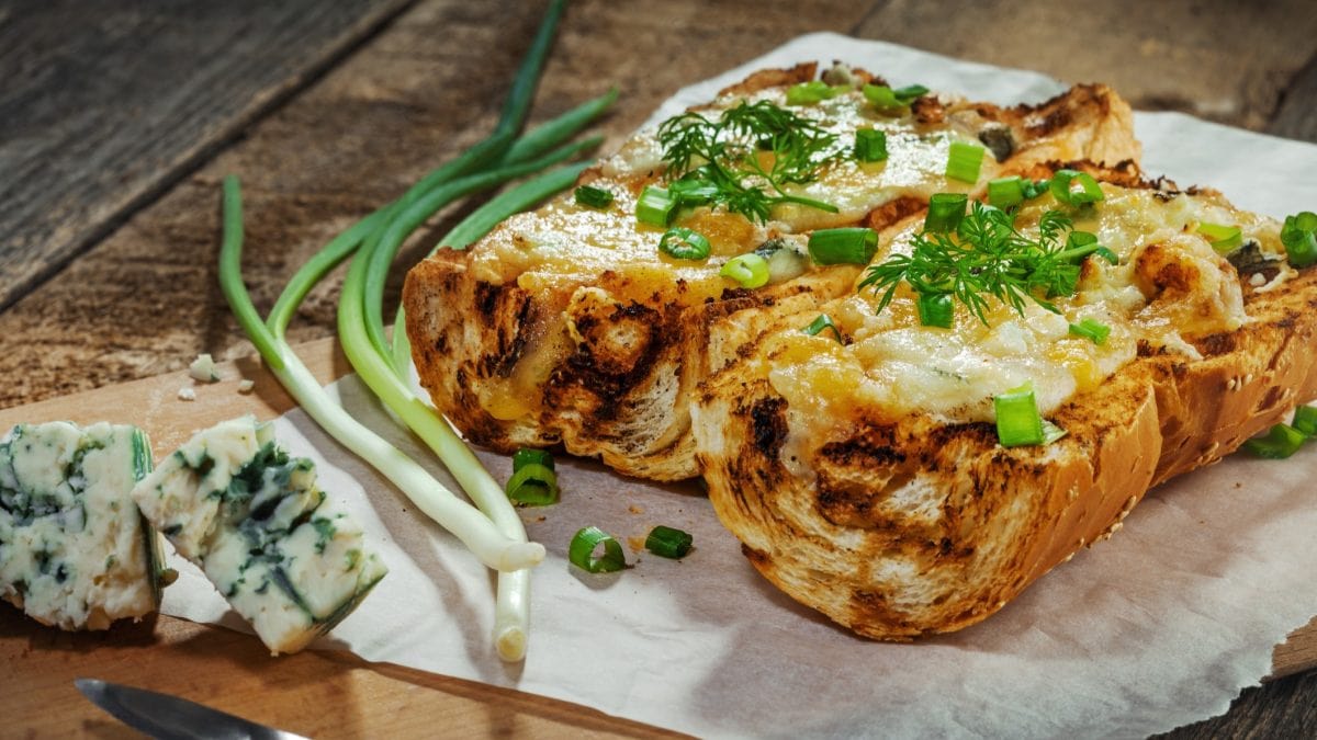 Draufsicht, schräg: Zwei halbierte Gorgonzola-Mozzarella-Baguette auf hellem Untergrund, der auf Holz liegt. Daneben liegen Blauschimmelkäsestücke sowie Frühlingszwiebeln und Kräuter.
