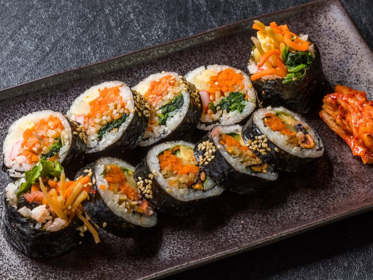 Kimbap auf einem schwarzen Teller serviert mit Kimchi. Der Hintergrund ist dunkelgrau.
