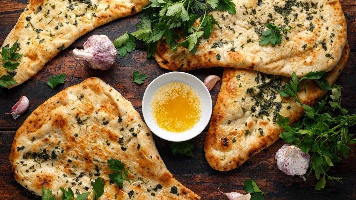 Knoblauch-Naan auf braunem Hintergrund verteilt. In der Mitte eine kleine Schüssel mit Knoblauchbutter. Knoblauch auf dem Bild verteilt.