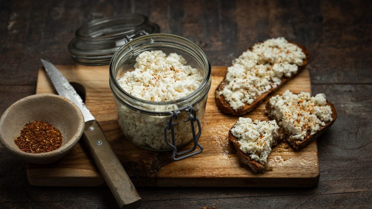 Ein dunkles Holzbrett auf dunklem Tisch mit einem Weckglas voll mit körnigem Frischkäse und beschmierten Broten. Daneben eine Schale rotes Gewürz und ein Messer. Alles von schräg oben fotografiert.