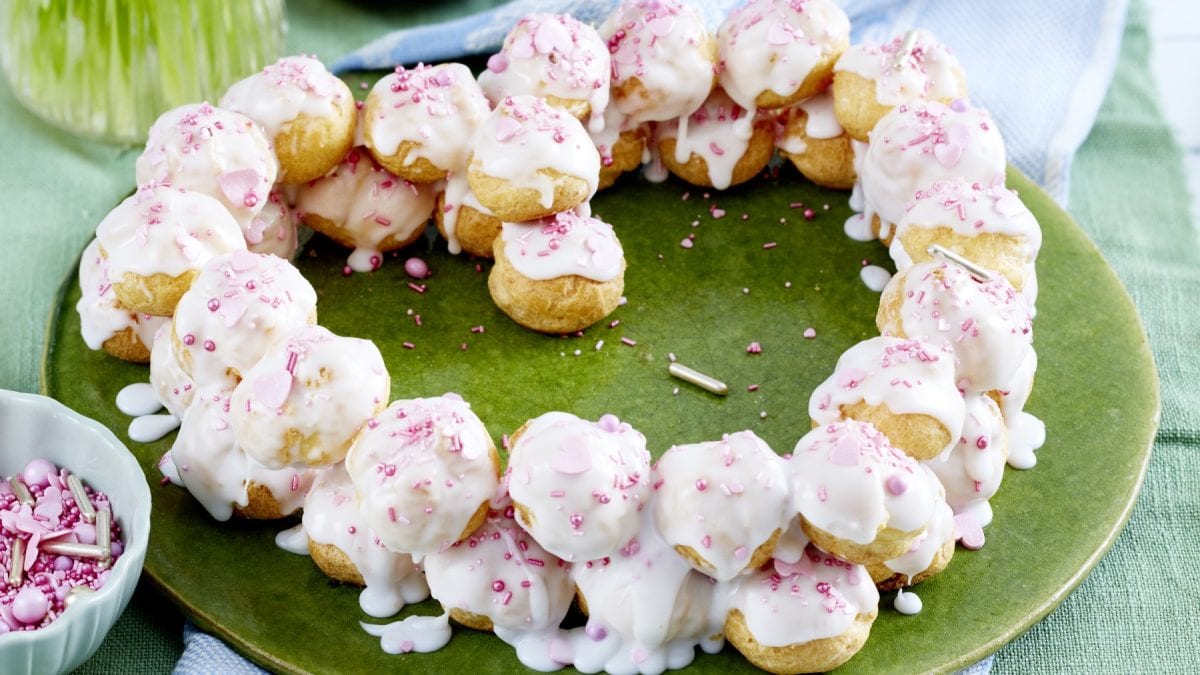 Ein Profiterol-Herz auf einem grünen Teller auf grünem Hintergrund. Links davon eine kleine Schüssel mit Zuckerstreusel.