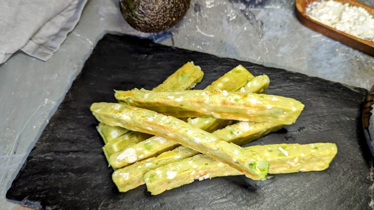 Mehrere Avocado-Feta-Stangen auf einer schwarzen Schieferplatte und grauem Steinuntergrund. Alles von schräg oben fotografiert.