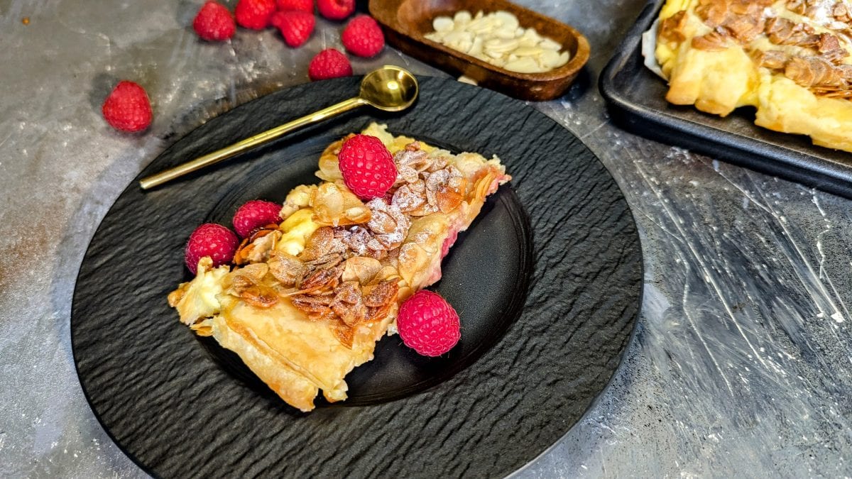 Eine Bienenstich-Schnitte mit Himbeeren auf einem schwarzen Teller mit goldenem kleinen Löffel auf grauem Steinuntergrund. Alles von schräg oben fotografiert.