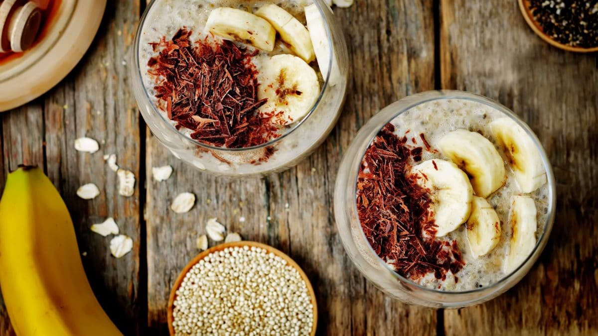 Zwei Gläser mit Quinoa-Pudding. Unten eine kleine Schüssel mit Quinoa und eine Banane. Der Hintergrund ist hölzern.