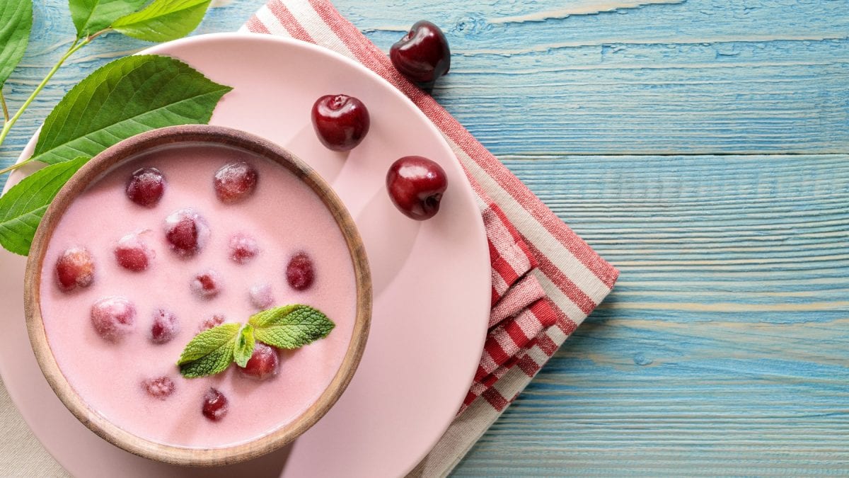Ungarische Sauerkirschsuppe in einer Schüssel, die auf einem rosa Teller und Geschirrtuch steht. Daneben einzelne Kirschen.