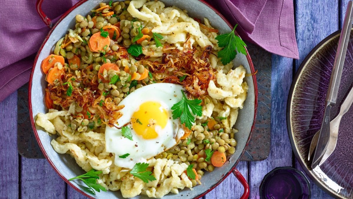 Draufsicht: Spätzle-Linsenpfanne mit Spiegeleiern in einer Pfanne auf blauem Holzuntergrund.