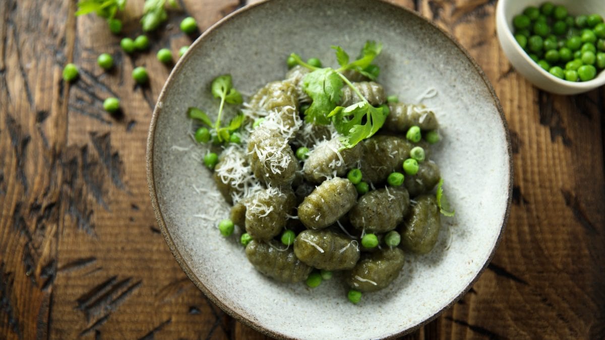Spinat-Gnocchi auf einem grauen Teller auf hölzernem Hintergrund. Auf dem Hintergrund Erbsen verteilt und rechts eine kleine Schüssel mit Erbsen.
