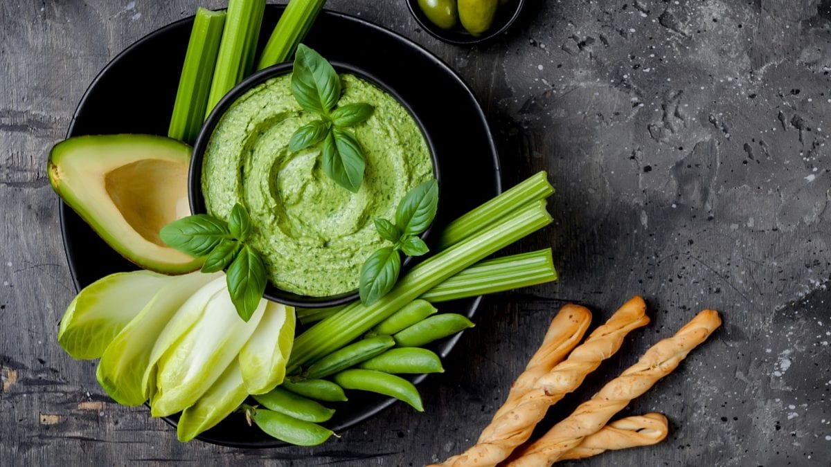Ein schwarzer Teller mit dem Staudensellerie-Pesto und grünem Gemüse direkt von oben auf dunklem Untergrund fotografiert.