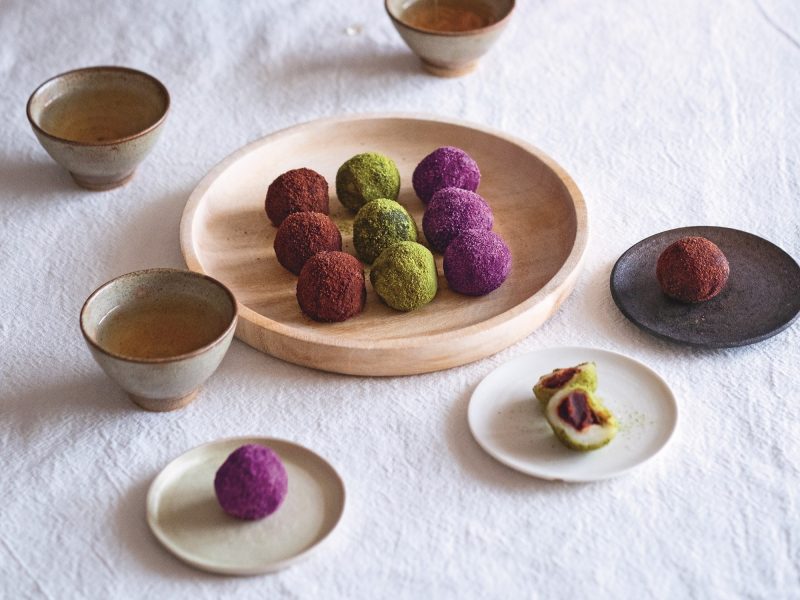Mehrere bunte Reiskugeln auf einem Tisch mit heller Tischdecke und einigen Tassen Tee. Alles von schräg oben fotografiert.