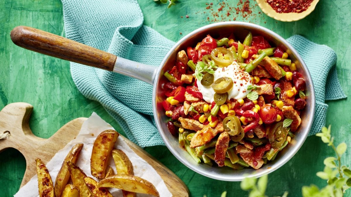 Tex-Mex-Pfanne vor einem türkisen Hintergrund auf einem blauen Geschirrtuch. Daneben ein Holzbrett mit Kartoffelspalten.