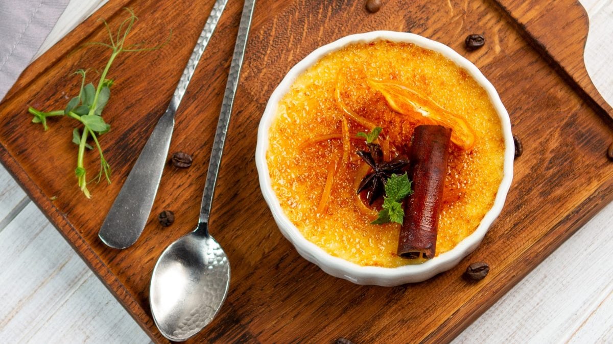 Thai-Tee Crème Brûlée in einer weißen Bowl auf einem braunen Schneidebrett. Links davon Besteck. Der Hintergrund ist hellgrau.
