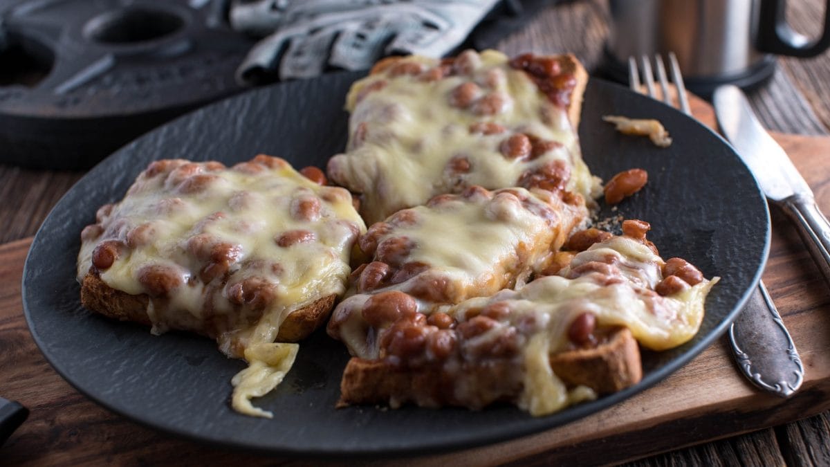 Draufsicht: Ein grauer Teller mit drei Broten. Es handelt sich um überbackener Baked-Beans-Toast, die mit Reibekäse bestreut sind.