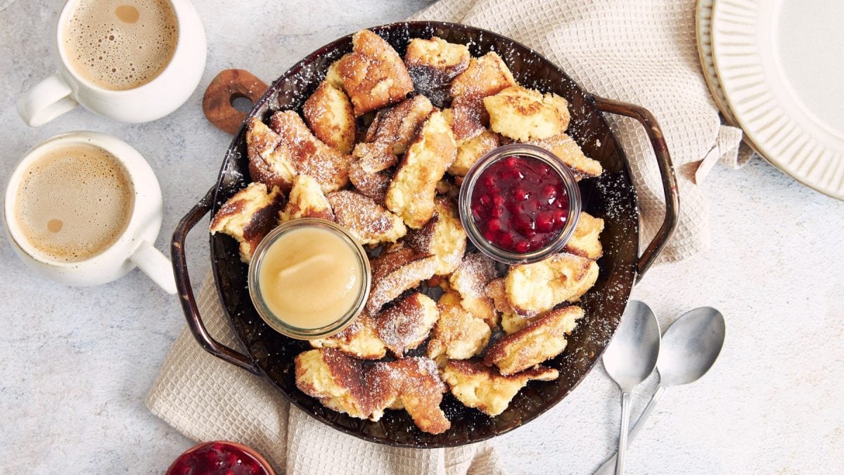 Veganer Kaiserschmarrn in einer schwarzen Pfanne mit Preiselbeermarmelade und Apfelmus serviert. Die Pfanne steht auf einem beigen Geschirrtuch und der Hintergrund ist hellgrau. Links auf dem Bild zwei Kaffeetassen, rechts zwei Löffel.