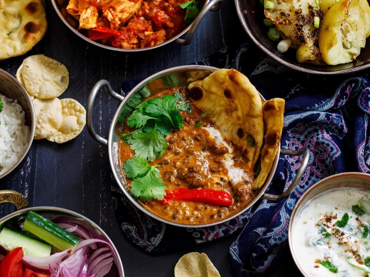 Indisches Curry mit Sahne, frischem Koriander und Naanbrot in einer traditionellen Metallschale, umgeben von Beilagen.