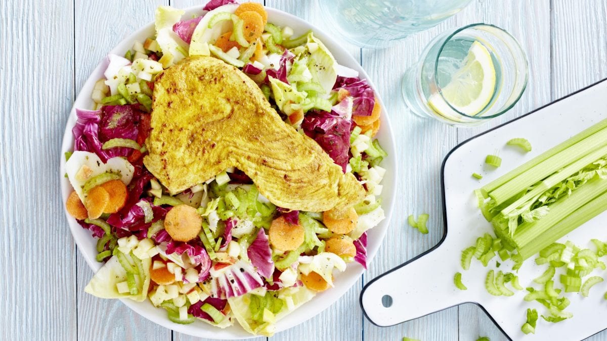 Wintersalat mit Putenschnitzel auf einem weißen Teller. Rechts davon ein Schneidebrett mit Staudensellerie und rechts oben auf dem Bild ein Glas Wasser mit Zitrone.