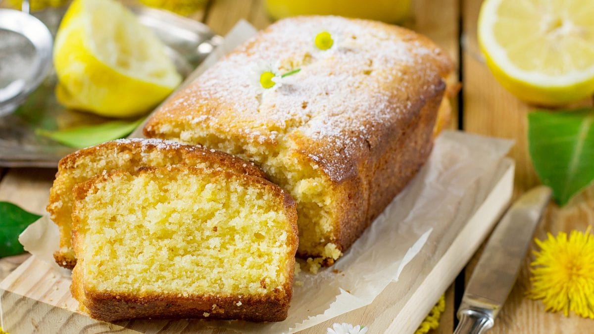 Der angeschnittene Zitronen-Kastenkuchen auf einem Holzbrett und rustikalem Holztisch. Daneben und dahinter gelbe Blüten und Zitronen. Alles schräg von oben und leicht von der Seite fotografiert.