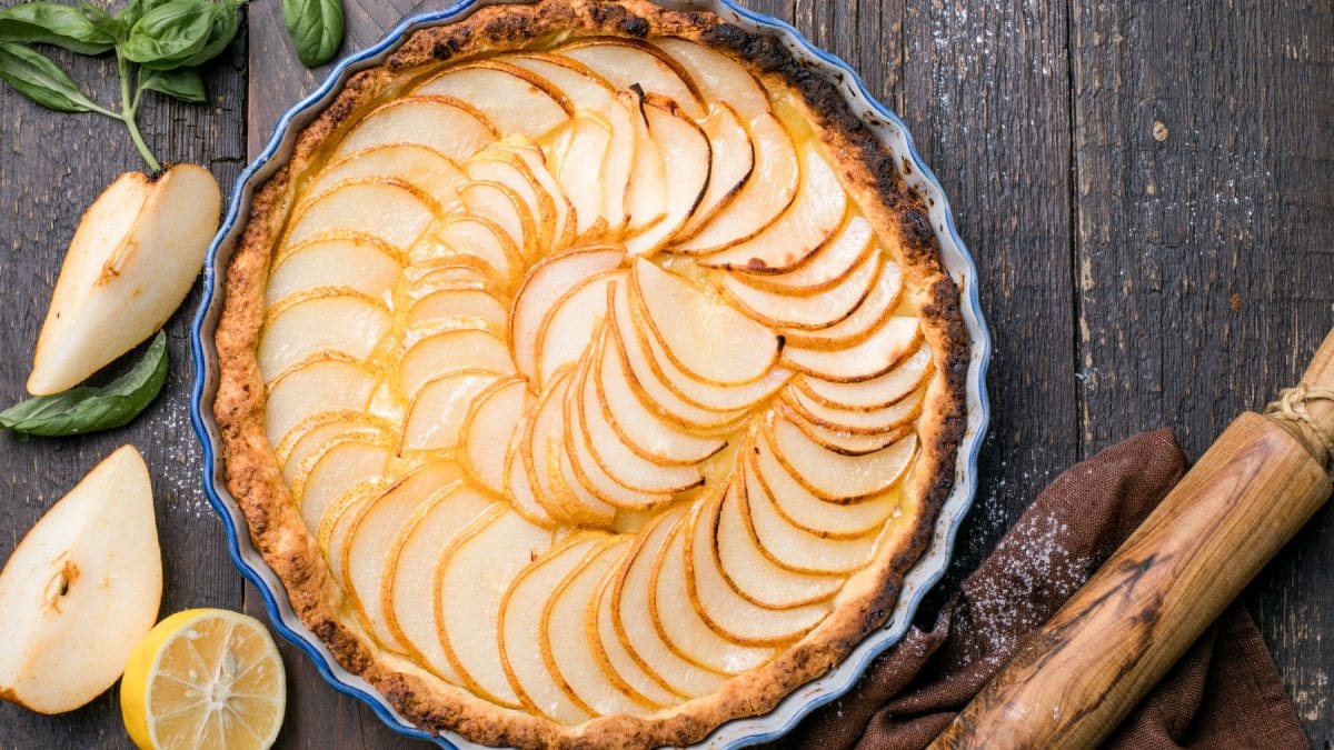 Draufsicht: Runde Zitronen-Tarte mit Birne auf Holzunterhgrund, drum herum Zitronen, Birnen und ein Nudelholz.