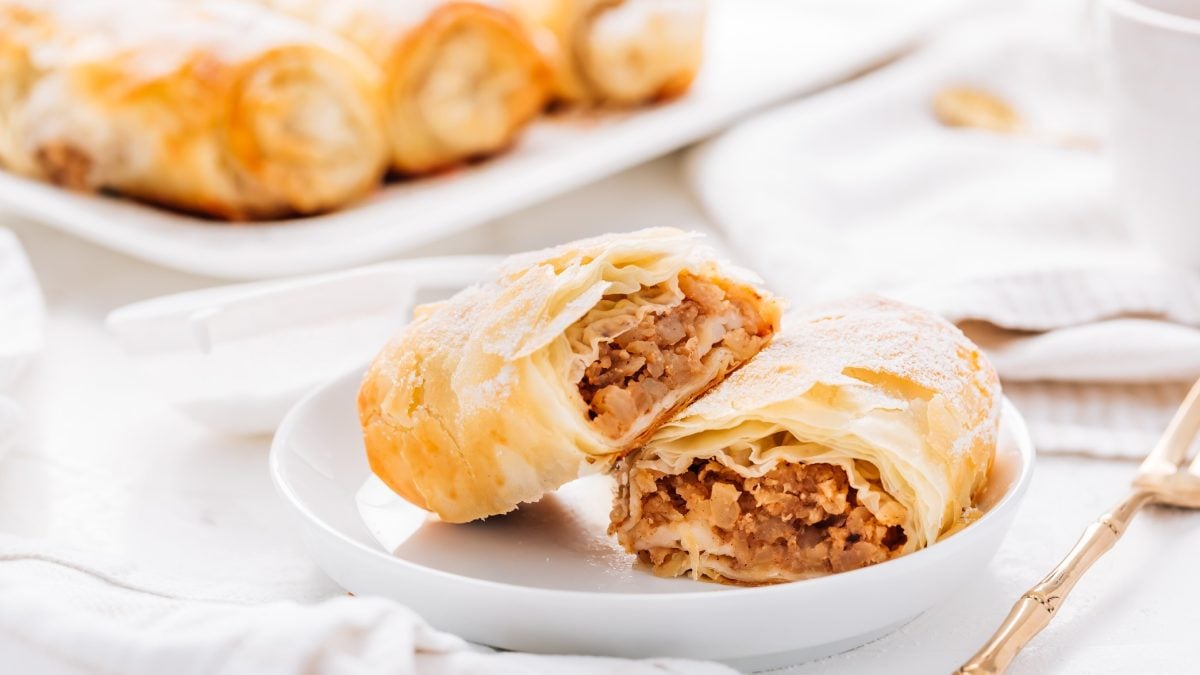 Ein weißer Teller mit einem halbierten Apfelstrudel-Röllchen und drei Stück im Hintergrund. Alles auf weißem Tisch mit weißer Tischdecke und sehr nah von der Seite fotografiert.