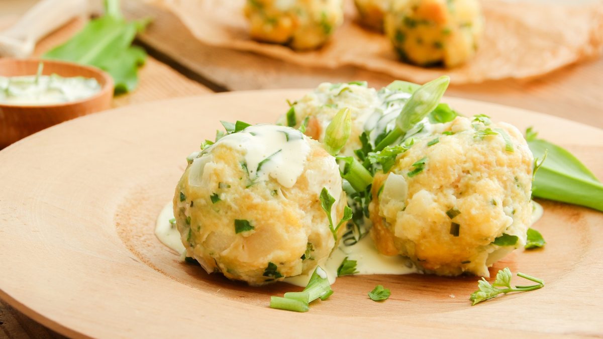Ein heller Teller mit drei Knödeln und der Kräuter-Buttermilch auf einem Holztisch. Im Hintergrund unscharf weitere Knödel erkennbar. Alles nah und von der Seite fotografiert.