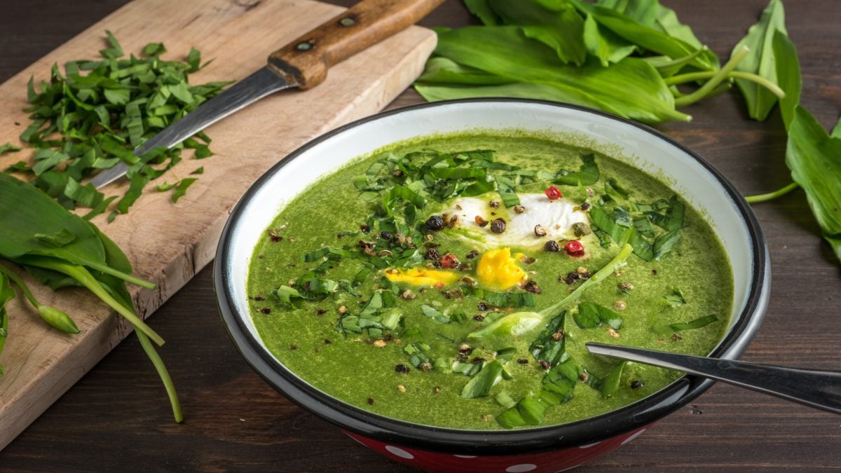 Die Bärlauchsuppe in einer hellen Schale auf einem Holztisch mit frischem Bärlauch im Hintergrund. Alles leicht oben von der Seite fotografiert.