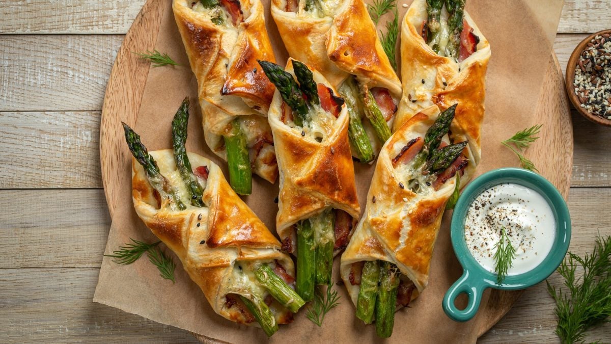 Blätterteigtaschen mit grünem Spargel und Bacon auf einem runden Holzbrett. Daneben ein weißer Dip.