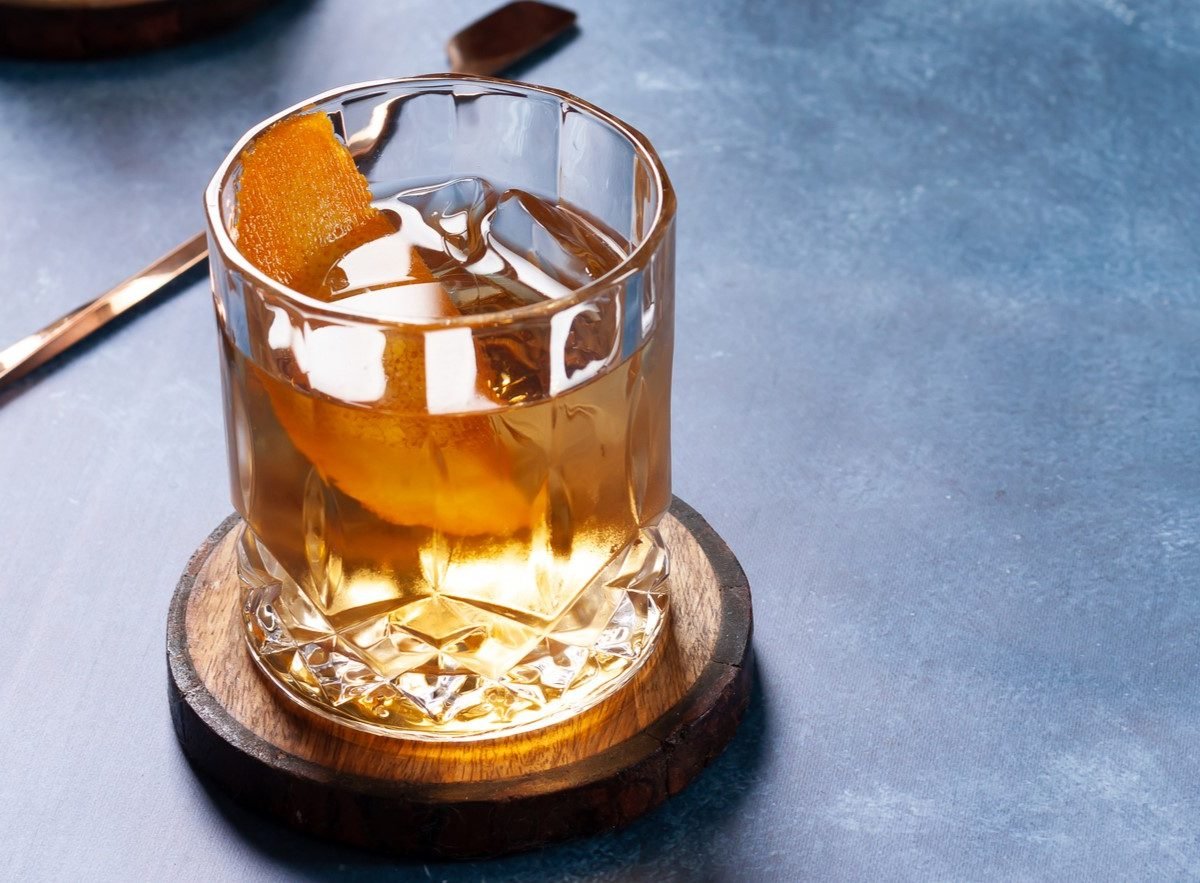 Zum Tag des Bourbon: Was ist Bourbon? Ein Glas mit Bourbon auf einem dunkelblauen Tisch mit Barlöffel. Frontalansicht.