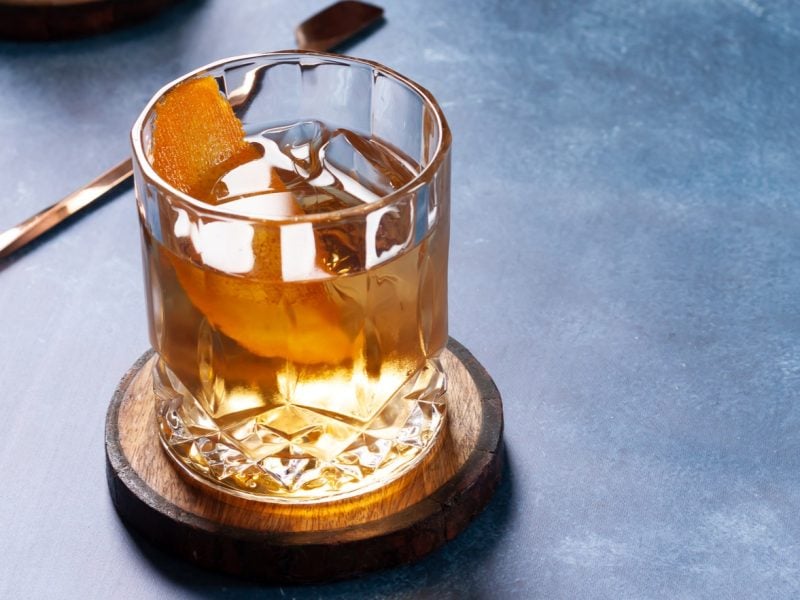 Zum Tag des Bourbon: Was ist Bourbon? Ein Glas mit Bourbon auf einem dunkelblauen Tisch mit Barlöffel. Frontalansicht.