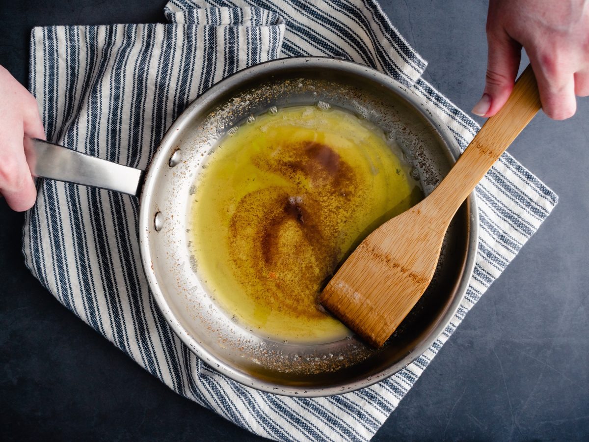 Braune Butter in einer Edelstahlpfanne von oben. Daneben eine Hand, die einen Holzkochlöffel hält.