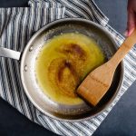 Braune Butter in einer Edelstahlpfanne von oben. Daneben eine Hand, die einen Holzkochlöffel hält.