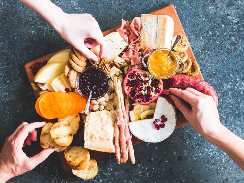 Ein stilvolles Charcuterie Board mit Käse, Feigen, Nüssen, Granatapfelkernen und Brot, umgeben von greifenden Händen.