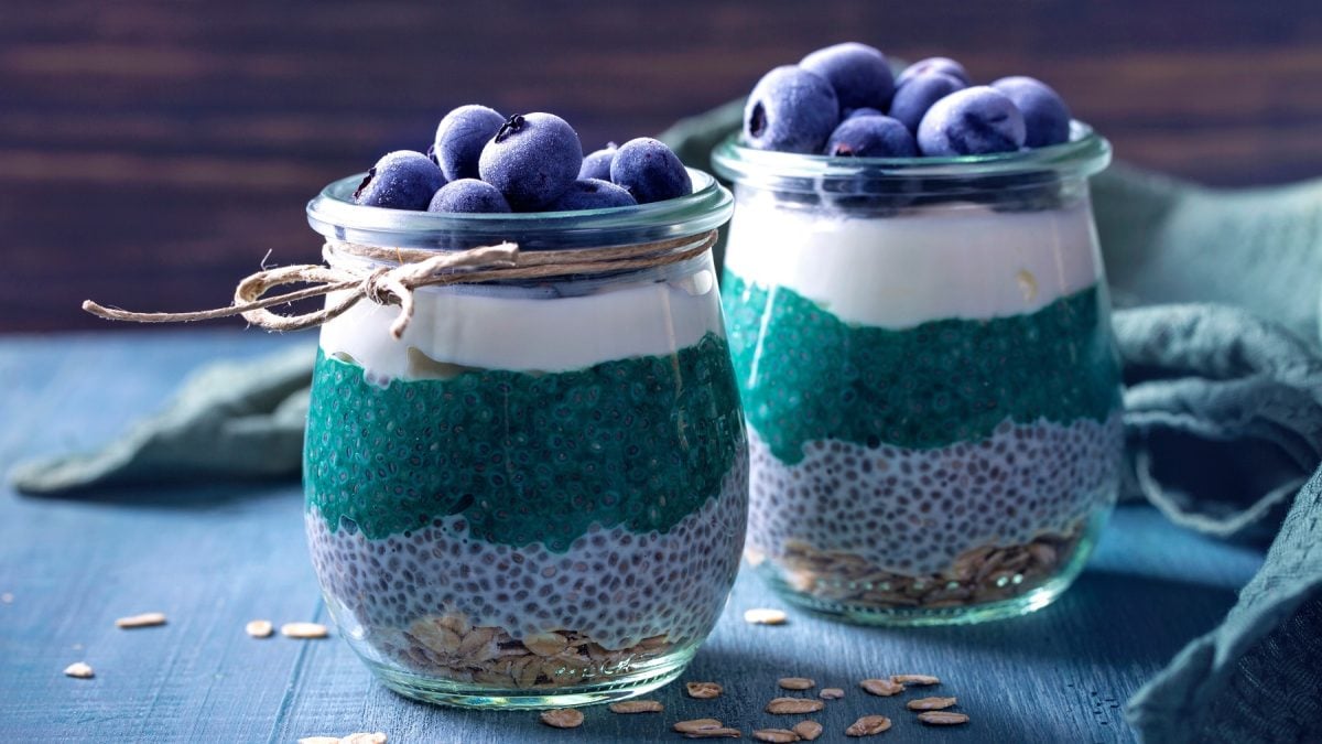 Zwei Gläser mit dem Chia-pudding auf einem blauem Tishc udn dunklem Holzhintergrund. Alles von der Seite fotografiert.