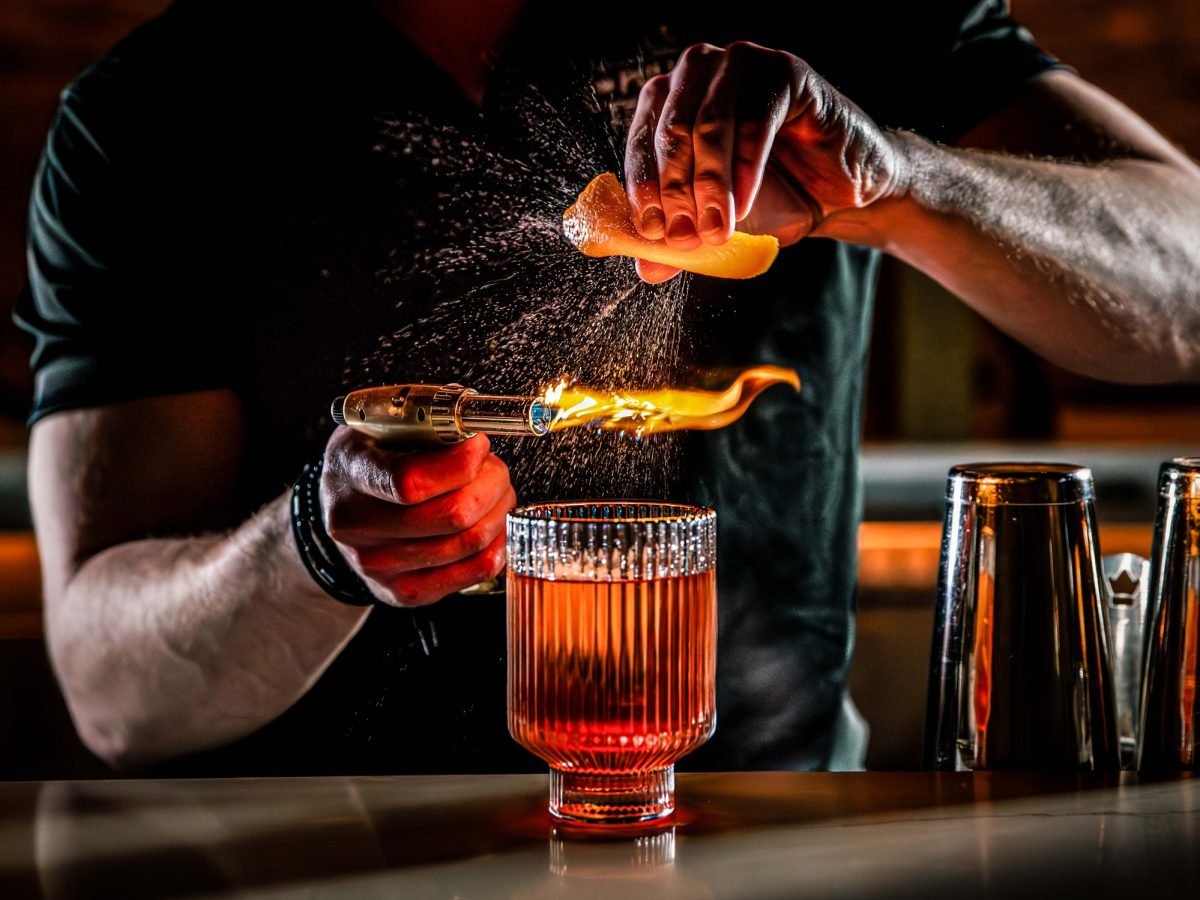 Ein Barkeeper mixt gerade einen Drink und dekoriert ihn mit Orange und Bunsenbrenner.