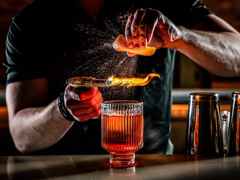 Ein Barkeeper mixt gerade einen Drink und dekoriert ihn mit Orange und Bunsenbrenner.
