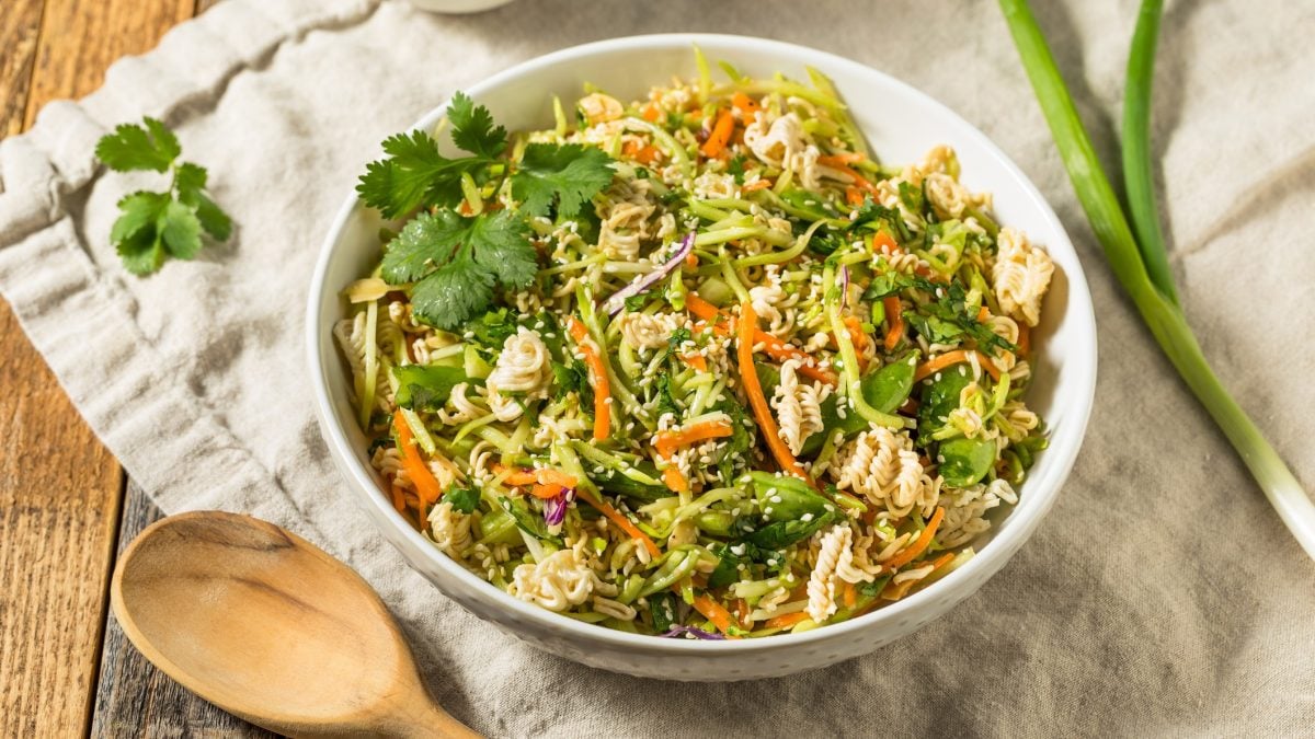 Eine weiße Schale mit dem crunchy Nudelsalat auf einem Holztisch mit cremefarbener Tischdecke. Alles von oben fotografiert.