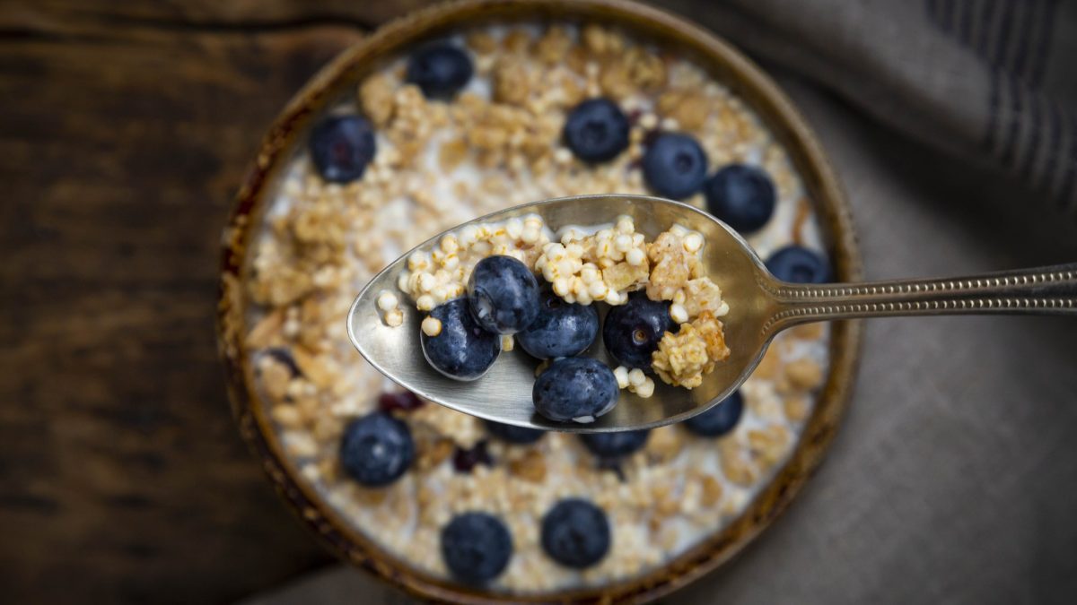 Draufsicht: Eine runde Beeren-Quinoa-Bowl steht auf einem Holztisch. Über der Bowl ist ein gefüllter Silberlöffel mit Beeren und Quinoa.