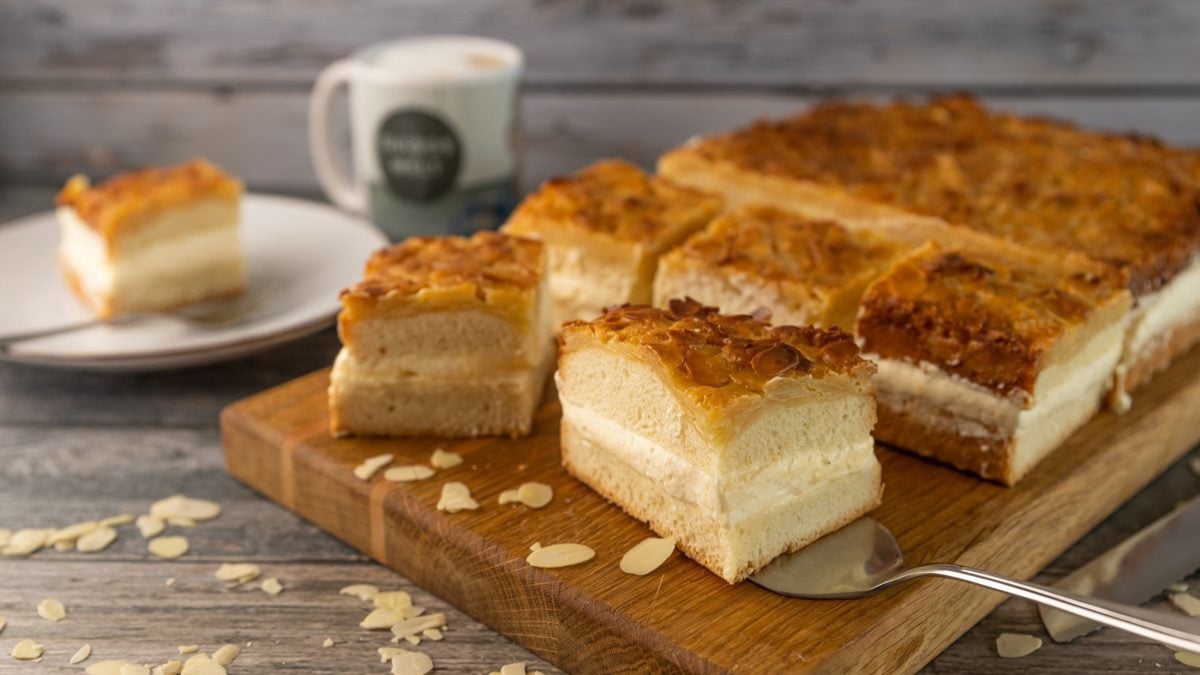 Frontal, leicht schräg von oben: Auf einem Holztisch liegt ein Holzbrett auf dem mehrere Stücke Bienenstich-Kuchen liegen. Im Hintergrund ist ein Teller mit einer Gabel und einem Stückchen Kuchen.