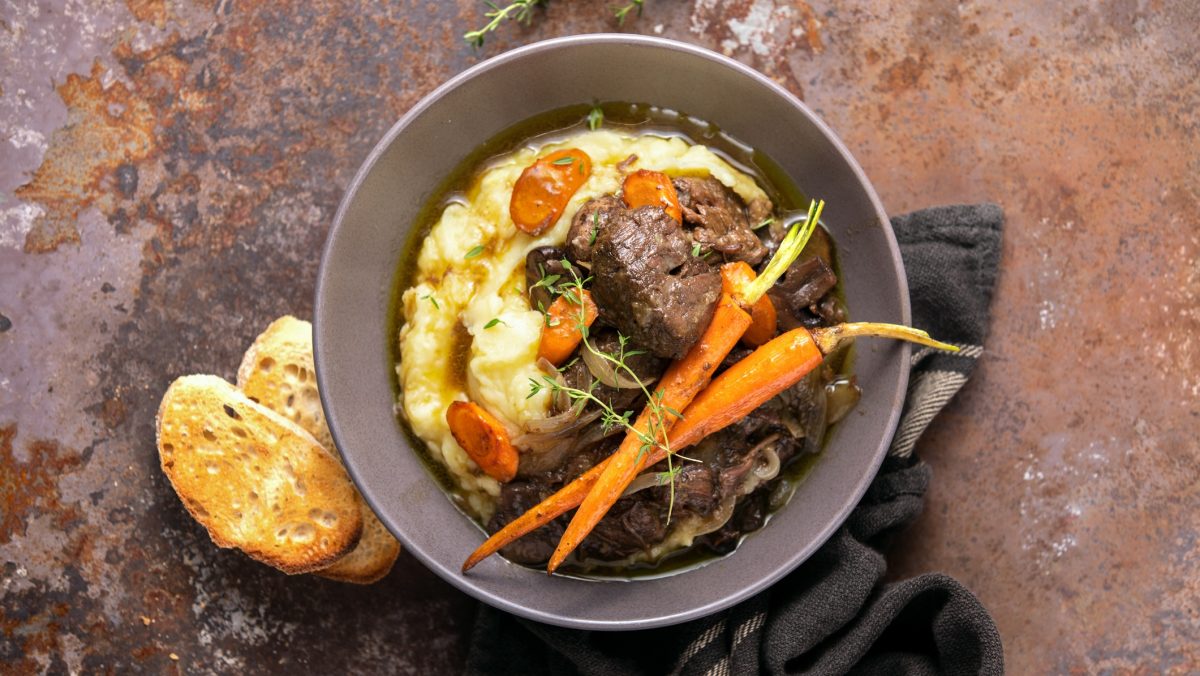 Draufsicht: Eine Ess-Schüssel Boeuf Bourguignon mit Kartoffelpüree steht auf einem rustikalen Untergrund aus Holz. Daneben liegt eine Scheibe geröstetes Weißbrot.