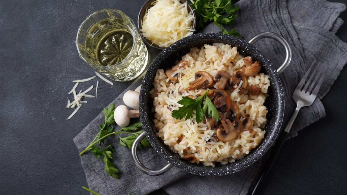 Draufsicht: Ein dunkler Topf Champignon-Risotto auf einem gräulichen Untergrund. Daneben sind Glas Weißwein, frische Pilze, frisch geriebener Käse und Petersilie.