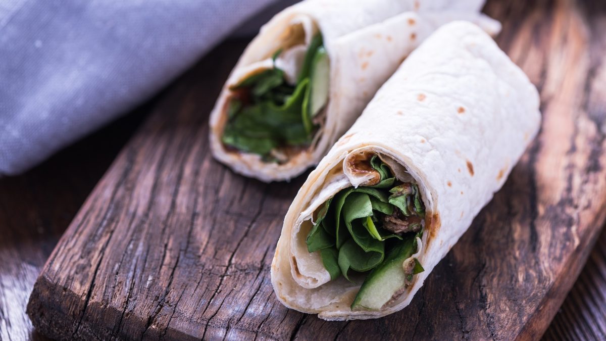Draufsicht, schräg: Zwei gerollte Dürüm Çiğ Köfte liegen auf einem dicken und schmalen Holzbrett. Aus den Wraps lugen Salatblätter heraus.