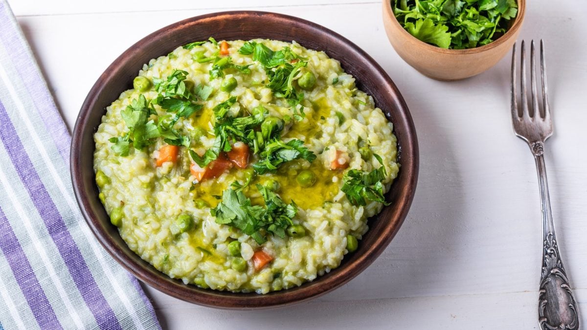 Draufsicht: Ein runder Teller Frühlings-Risotto mit Gartengemüse auf einem weißen Untergrund. Daneben liegt ein blau-weiß-gestreiftes Küchentuch. Darüber hinaus gibt es eine kleine Schale mit frischer Petersilie sowie eine Gabel.
