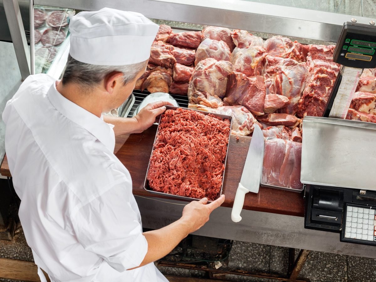 Draufsicht, schräg: Ein Metzger steht mit dem Rücken zu uns vor seiner Verkaufs-Theke. In der Hand hält er eine Form mit Hackfleisch. In der Auslage liegen viele verschiedene Fleischstücke.