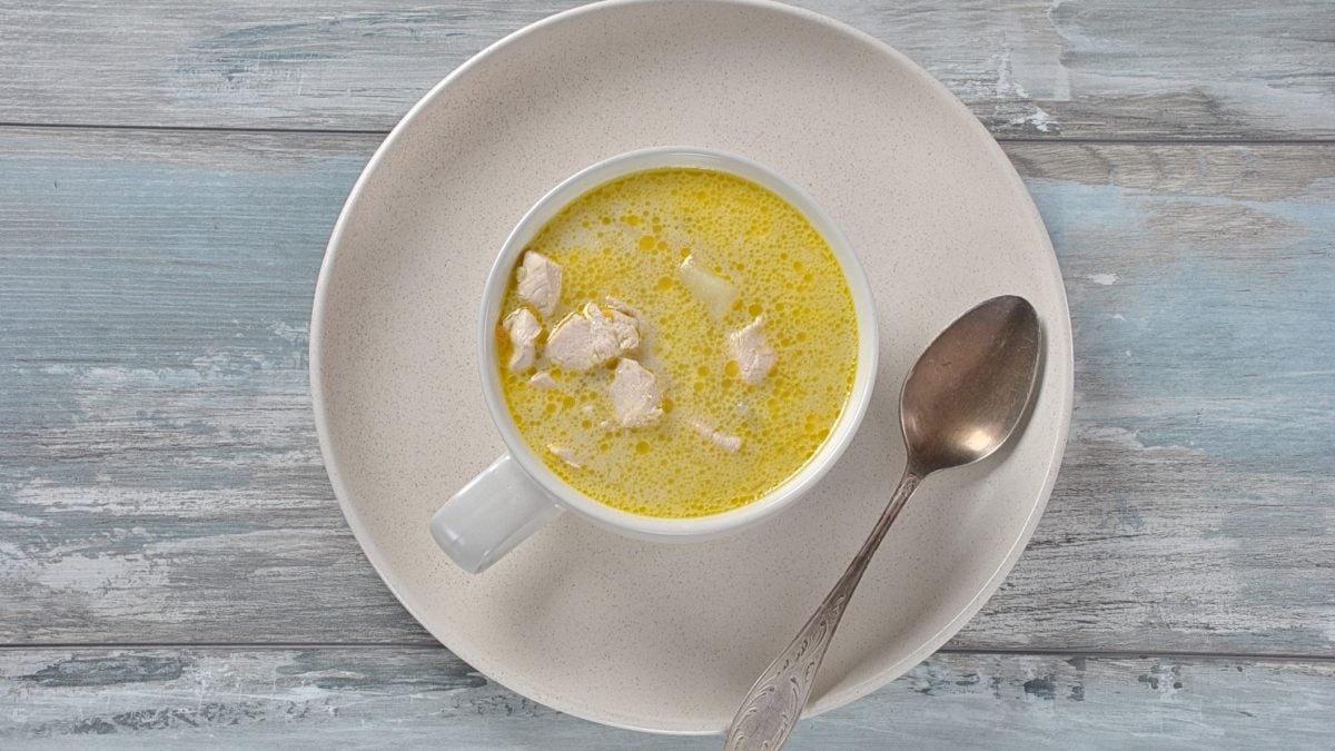 Draufsicht: Auf melierten, hellblauem Untergrund aus Holz lieget ein großer, runder und heller Teler. Auf steht eine gleichfarbige Tasse mit einer Kartoffel-Kokos-Suppe mit Hühnchen. Auf dem Teller liegt ein Suppenlöffel.