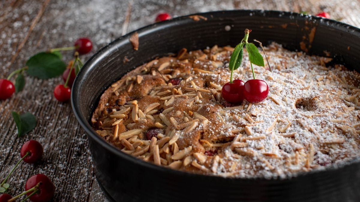 Draufsicht, schräg: Eine dunkle, runde Springform mit einem Kirschmichel liegt auf einem Untergrund aus Holz. Der Kuchen ist zur Hälfte mit Puderzucker bestreut.