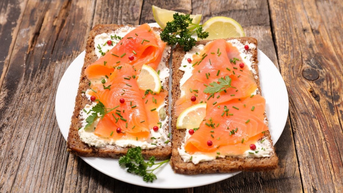 Draufsicht, schräg: zwei Lachs-Sandwich mit Hüttenkäse liegen parallel auf einem weißen Teller. Der liegt auf einem Untergrund aus Holzplanken.