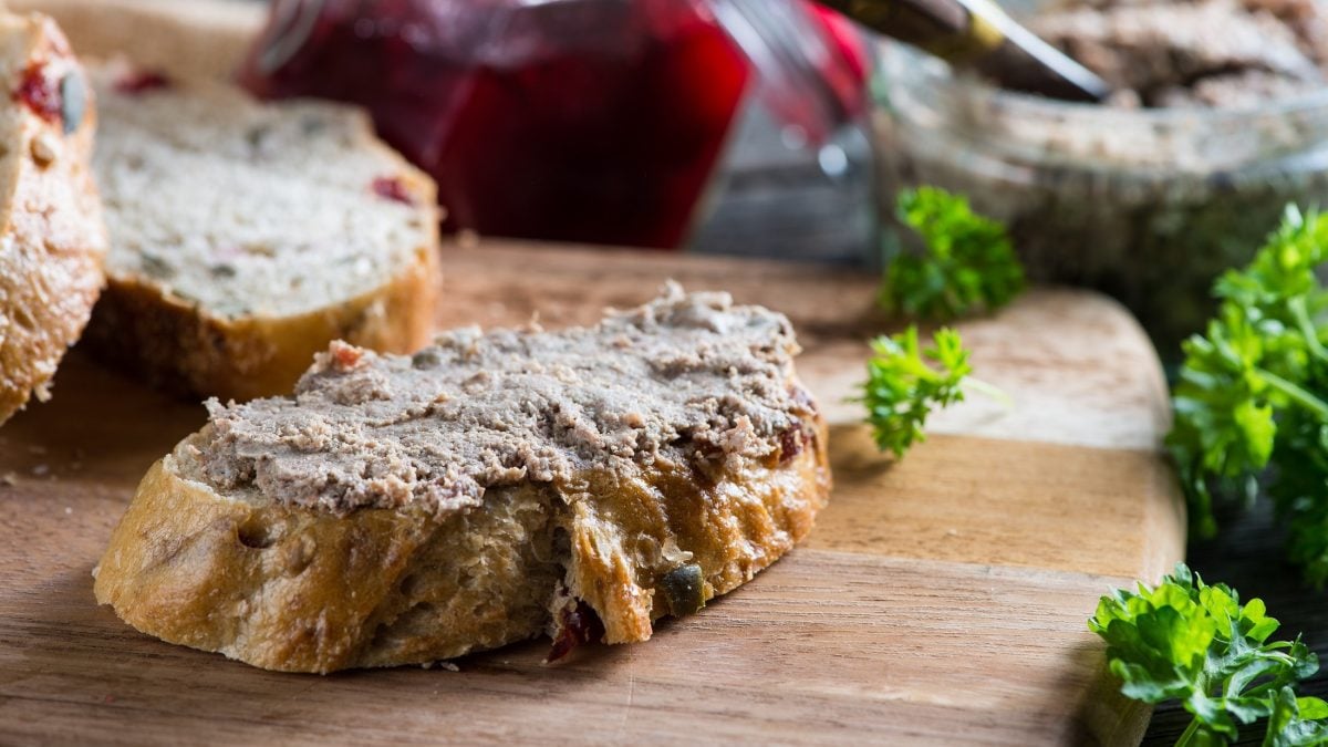 Draufsicht schräg: Auf einem Holzbrett liegt ein Weißbrot, dass mit Lamm-Leberwurst mit Masala bestrichen ist. Daneben liegen frische Petersilie und weitere Brotscheiben.