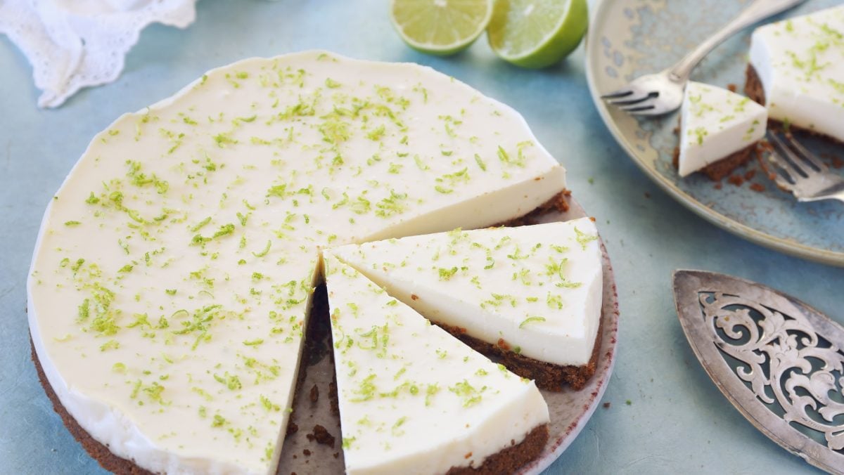Draufsicht: Ein runder Limetten-Kuchen steht auf einem Tisch mit einer hellblauen Tischdecke. Der Kuchen ist angeschnitten. Auch auf dem Bild: zwei Kuchenstücke, ein Kuchenheber aus Metall und eine weiße Tasse mit schwarzem Kaffee.