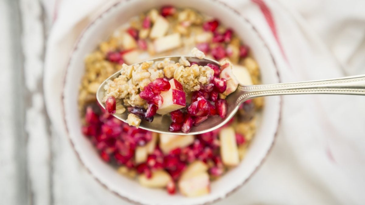 Draufsicht: Eine Schüssel Nuss-Granola mit Granatapfel. Darüber ist ein Löffel mit dem gleichen Granola.