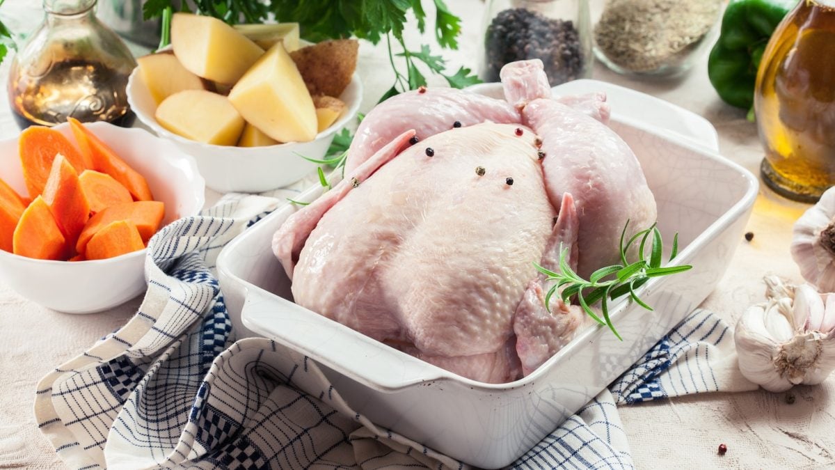 Draufsicht, schräg: Ein rohes Hähnchen liegt in einer Auflaufform. Drum herum stehen weiße kleine Schüsseln mit geschnittenem Gemüse, Kräuter, Öl und Gewürzen. Aus ihnen soll ein Ofenhähnchen mit Gemüse zubereitet werden.